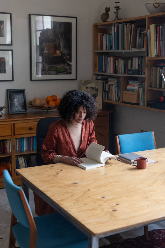 Dona asseguda llegint un llibre en una taula en una habitació plena de llibres i fotografies