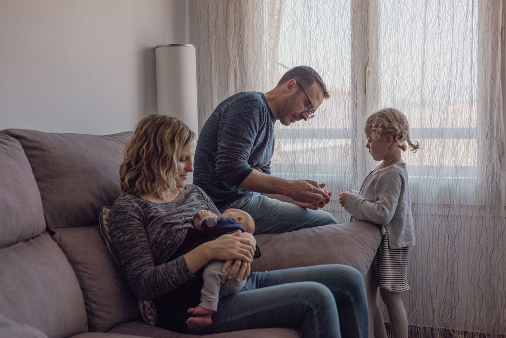 Fotografia de llum natural en família - mare, pare i filla a casa amb el nadó